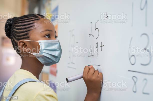 Portrait+of+african+girl+wearing+face+mask+and+writing+solution+of+sums+on+white+board+at+school.+Black+schoolgirl+solving+addition+sum+on+white+board+during+Covid-19+pandemic.+School+child+thinking+while+doing+mathematics+problem+and+wearing+surgical+mask+due+to+coronavirus.