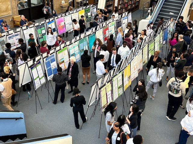 Students, parents, teachers, mentors and guests look at posters during the 2024 Research Practicum Symposium. 